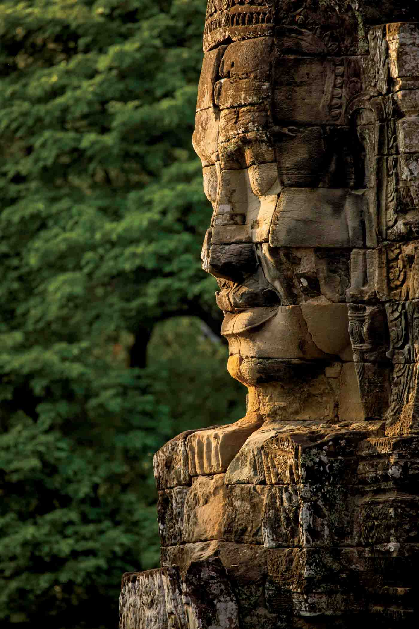 A Huge Face Sculpted In Stone, Watches Over Visitors To The Temple Of Bayon, Angkor, Cambodia, In The Background, A Wall Of Green Jungle Surrounds It