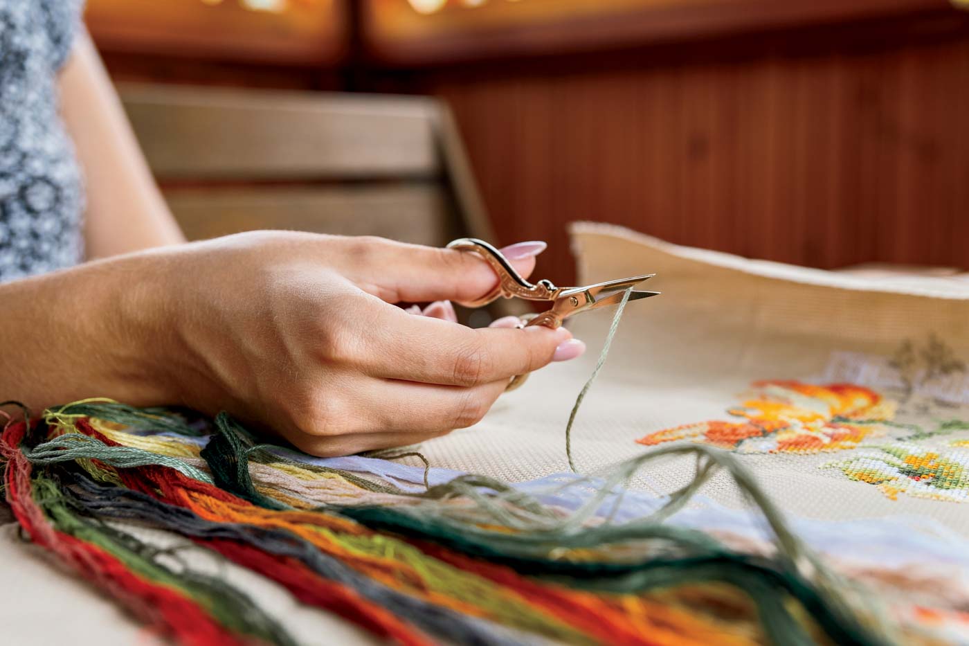 A Hand Holding Embroidery Scissors About To Cut A Thread. A Hand With Scissors Cuts The Thread. Close Up Of Cross Stitch. Crafts And Embroidery.